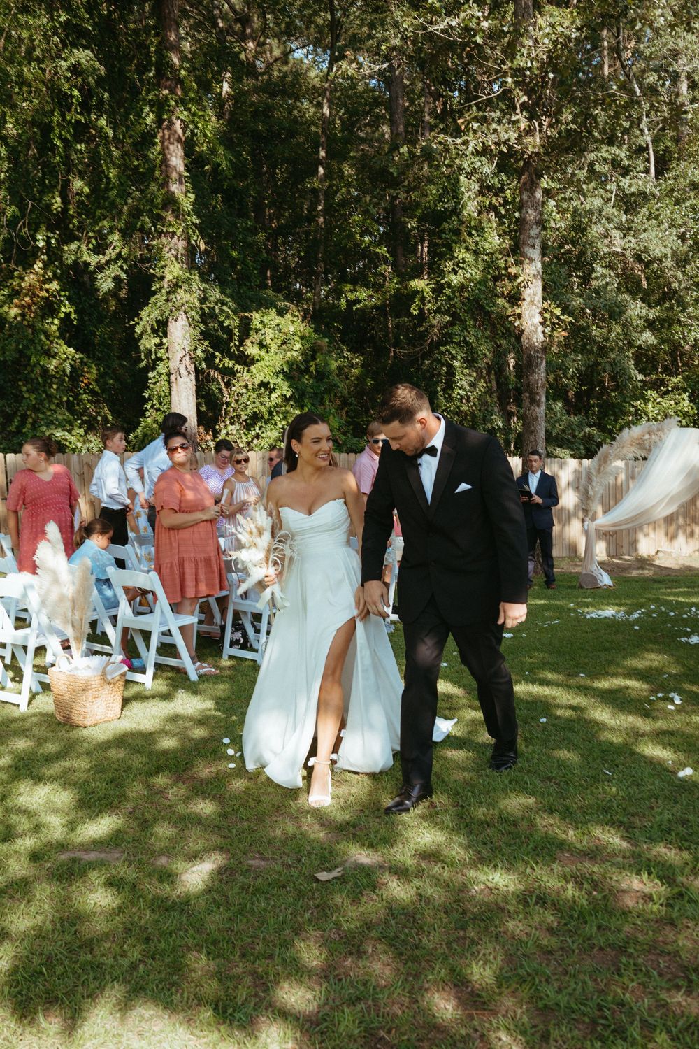 Bride and groom walk down outdoor wedding aisle surrounded by guests seated in white chairs among tall trees.
