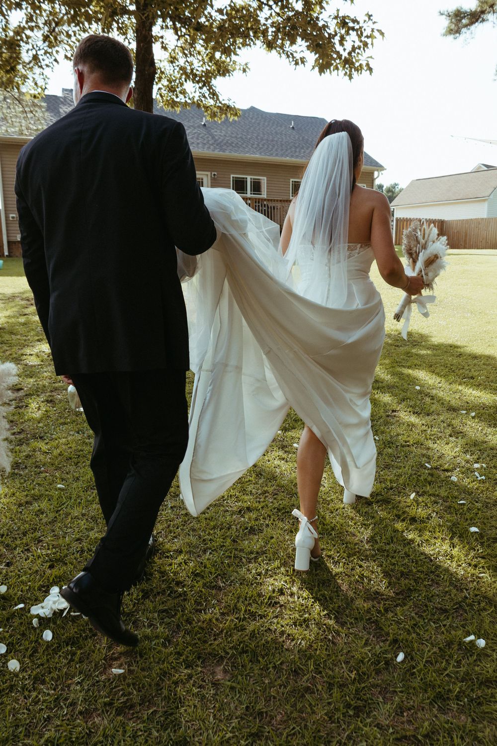 Groom lifts bride's white veil and train as couple walks across green lawn after outdoor wedding ceremony.