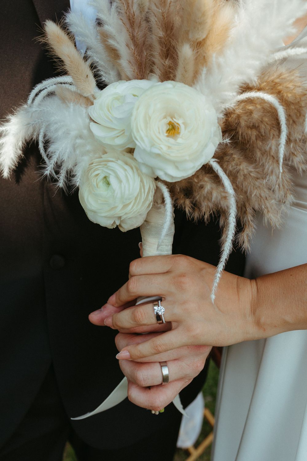 Bride and groom holding a boho wedding bouquet with white ranunculus and pampas grass, showing off wedding rings.