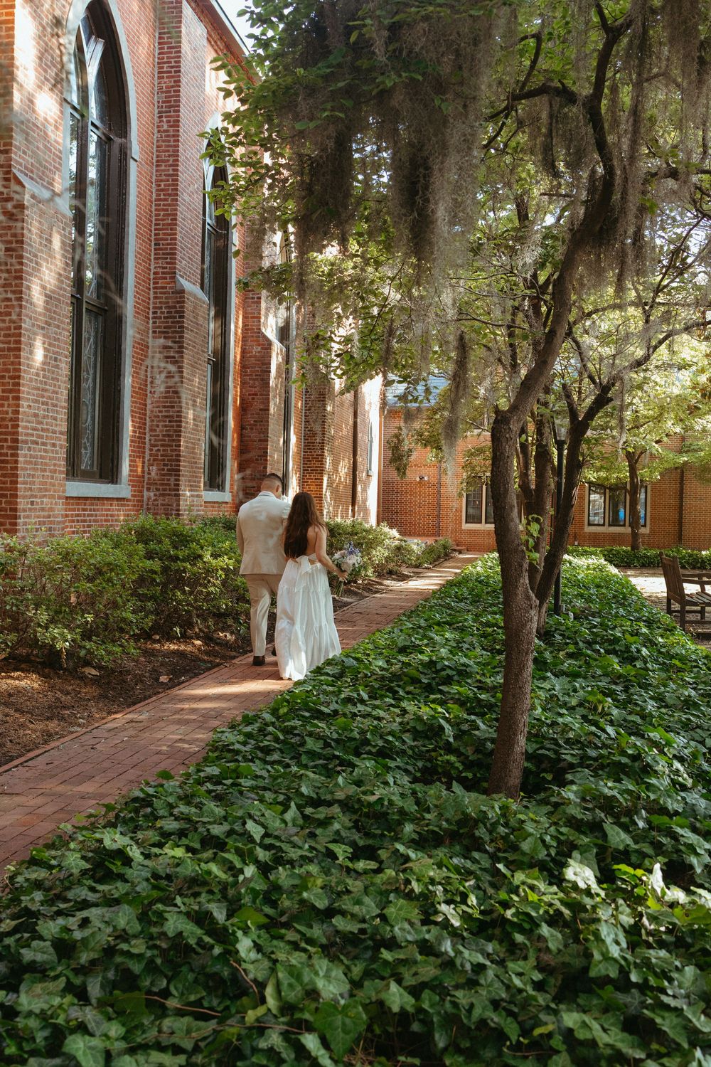 Bride and groom walking along a brick pathway beside a historic red brick church surrounded by lush ivy gardens.
