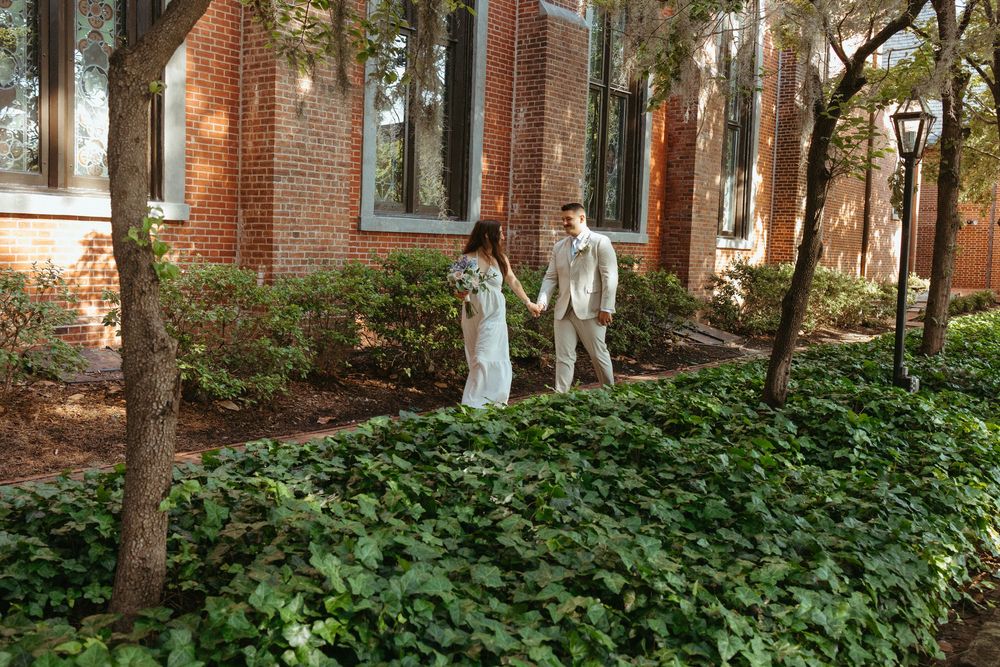 Bride and groom walk hand in hand through lush ivy garden beside brick building on their wedding day.