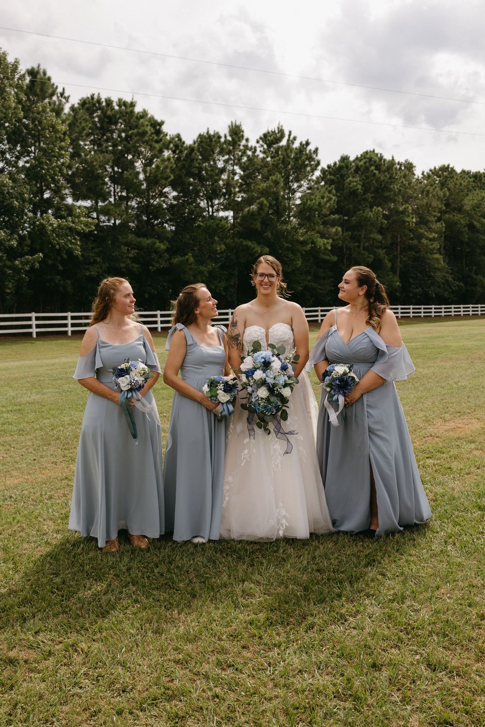 Bride in white gown with three bridesmaids in dusty blue dresses holding bouquets on a green lawn.
