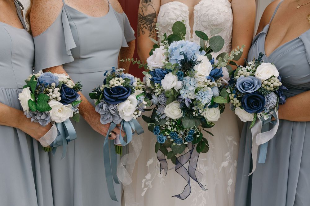 Bride in lace gown and bridesmaids in dusty blue dresses holding bouquets of navy, white, and blue flowers.