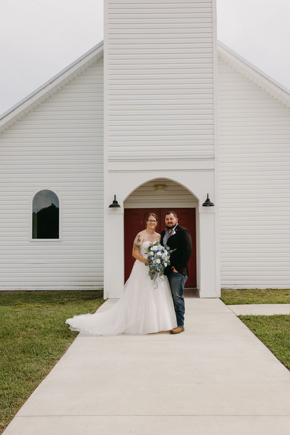 Bride and groom posing together in front of a white chapel with a red door on their wedding day.