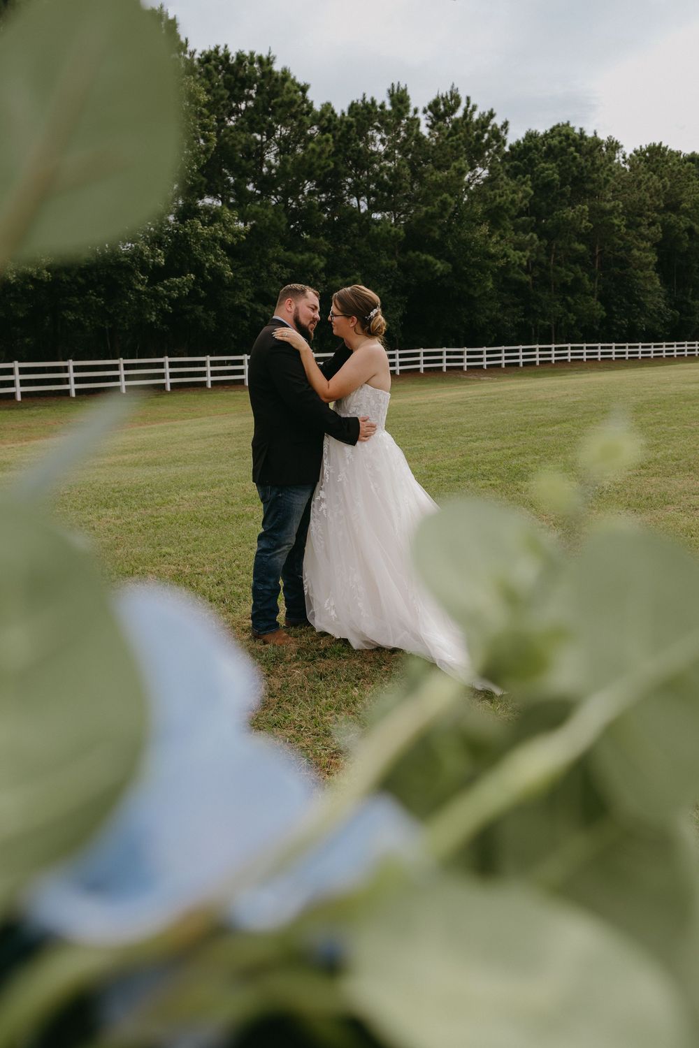 A bride and groom share a romantic moment on a green lawn with a white fence and trees in the background.