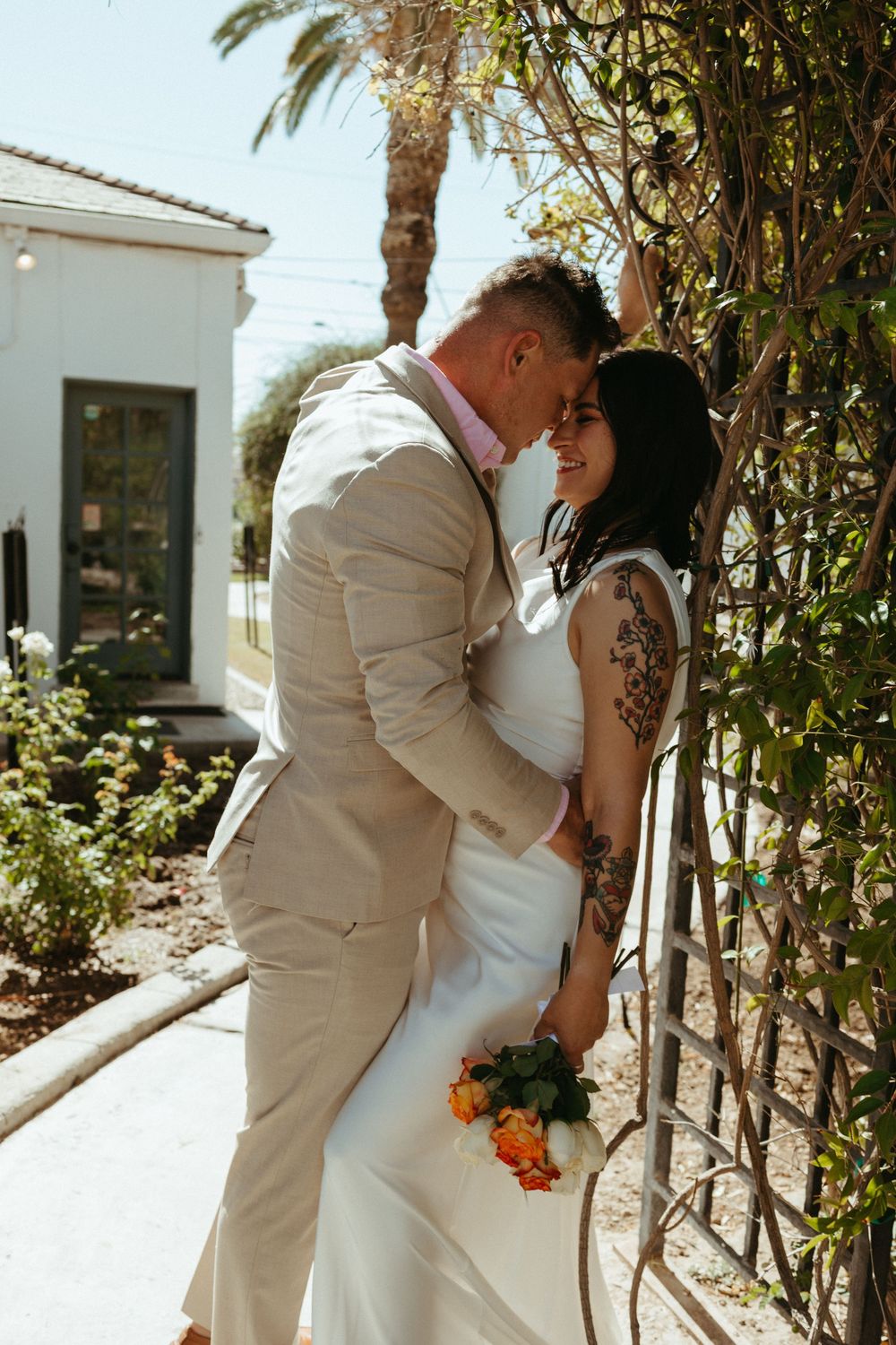 A bride and groom share a romantic moment outdoors, she in a white gown with floral tattoos, he in a tan suit.