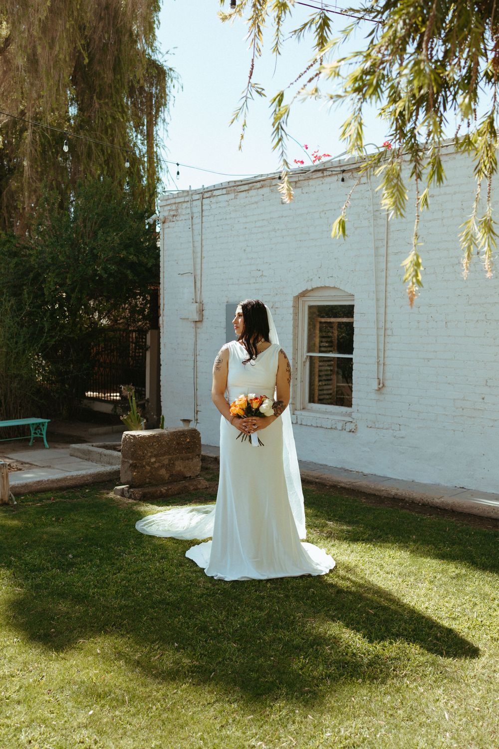 Bride in white gown holding colorful bouquet stands on green lawn beside white brick building on sunny day.