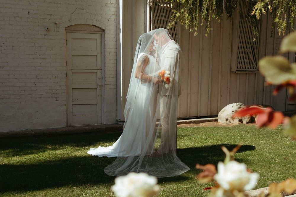 Bride and groom share a kiss under flowing white veil in outdoor garden wedding ceremony setting.