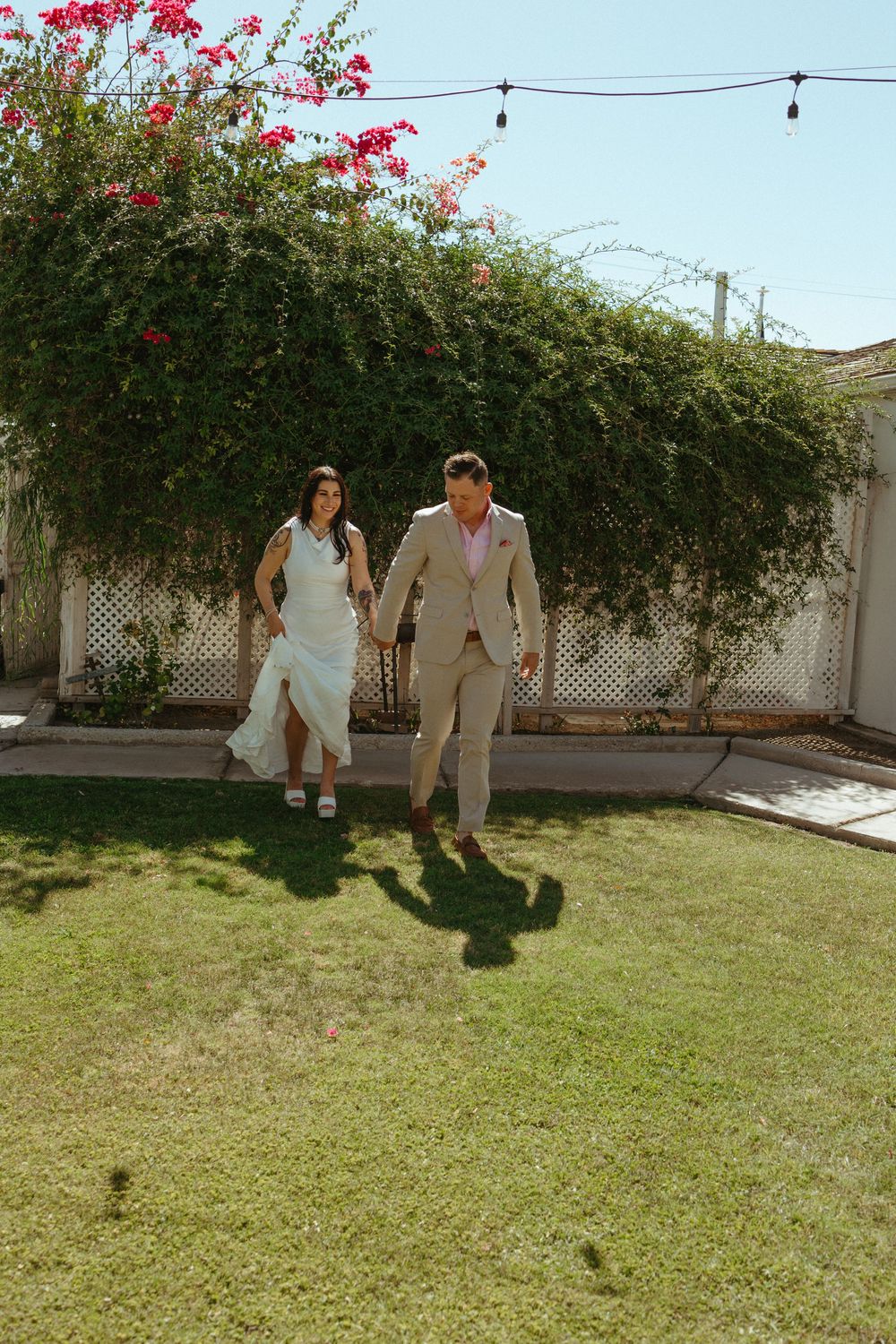 A couple in wedding attire holds hands while walking on green grass in a sunny backyard with blooming bougainvillea.