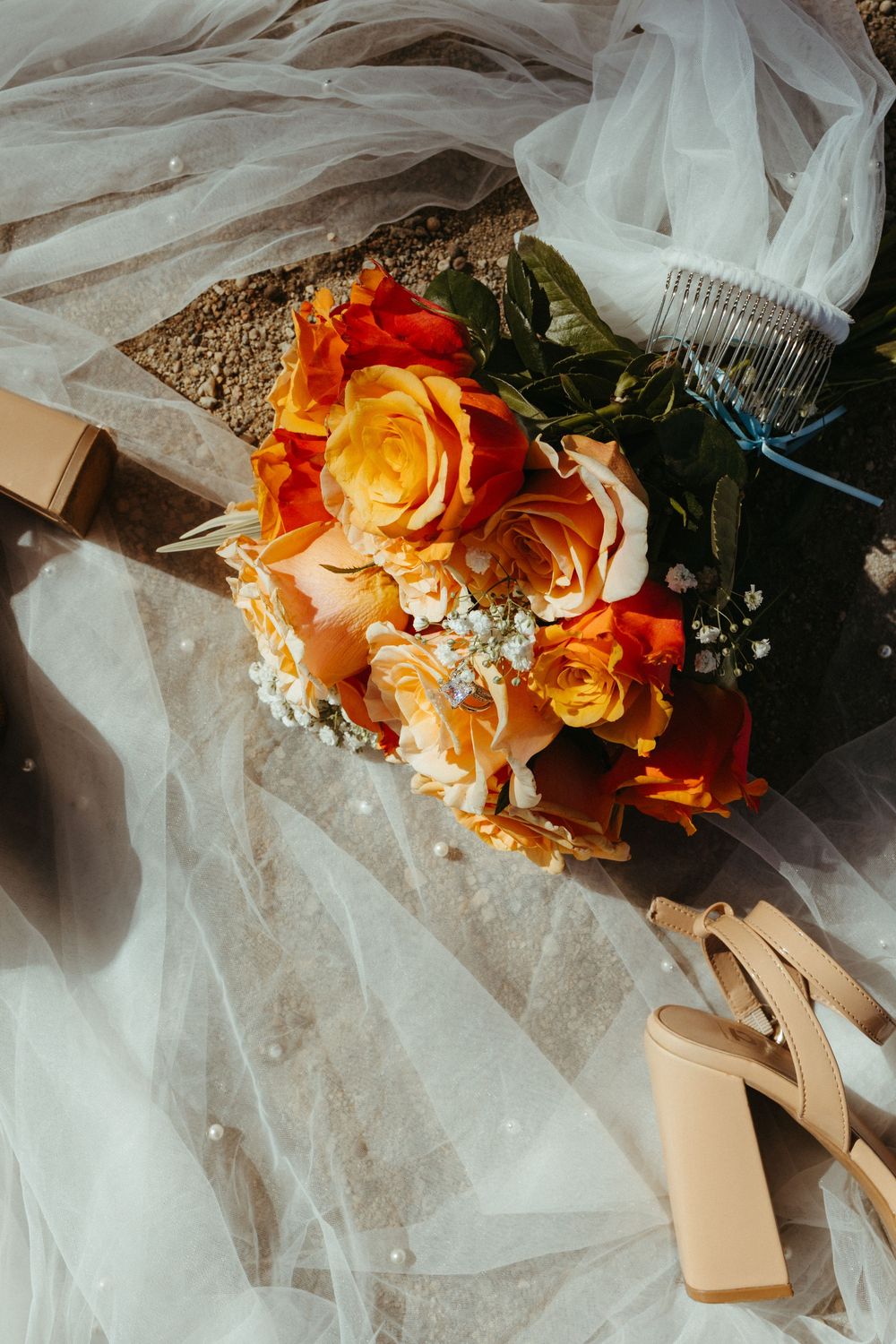 Bridal flat lay with orange and yellow rose bouquet, nude heels, pearl veil, and hair comb on white tulle.