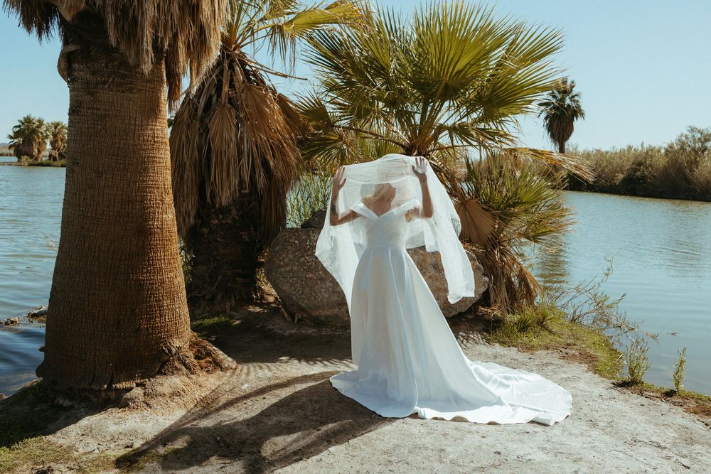 Bride in white gown holding veil by palm trees near a serene lake on a sunny day.