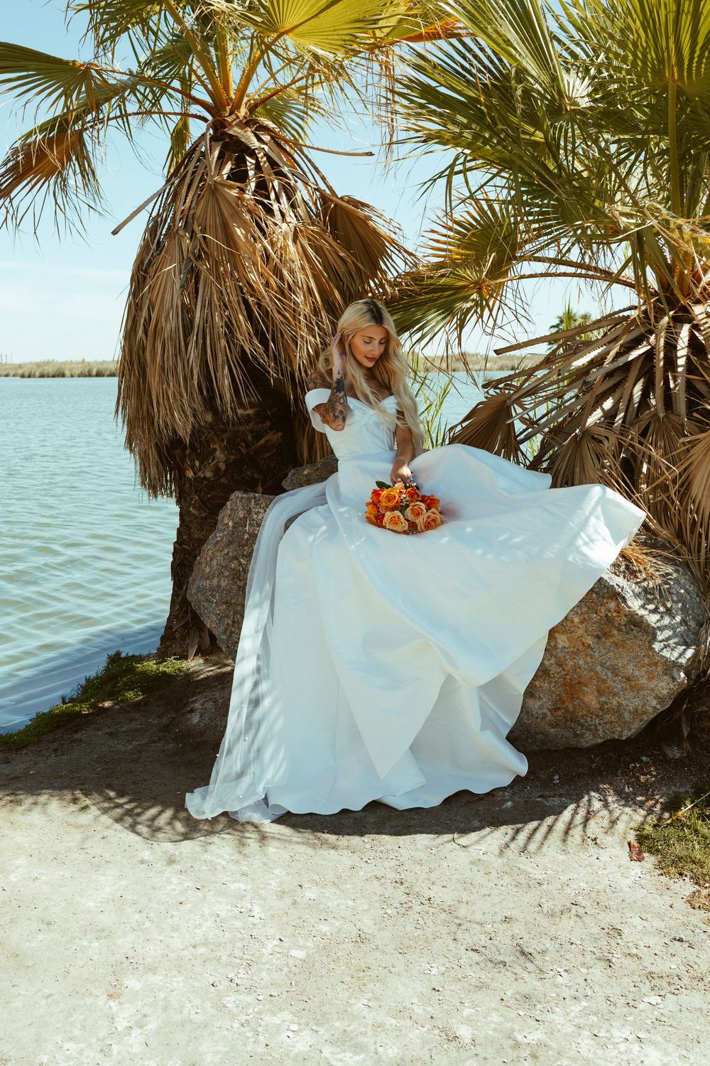 Bride in white ball gown sitting on rocks near palm trees by water, holding orange bouquet.