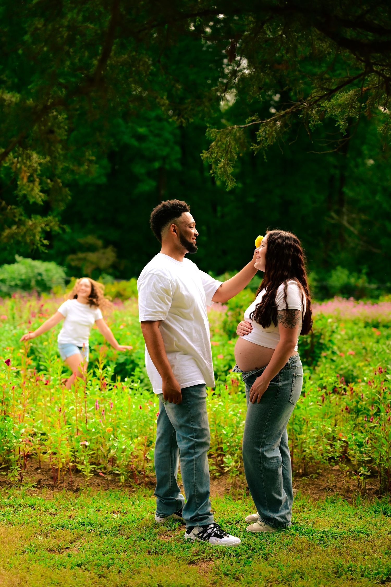 A man places a flower in a pregnant woman's hair in a lush garden, while a child runs joyfully in the background.