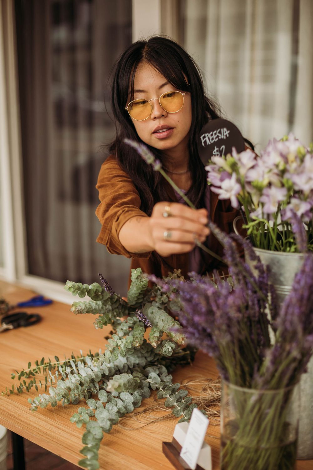 A person in glasses and brown jacket arranges purple and green flowers in a vase on a wooden table.
