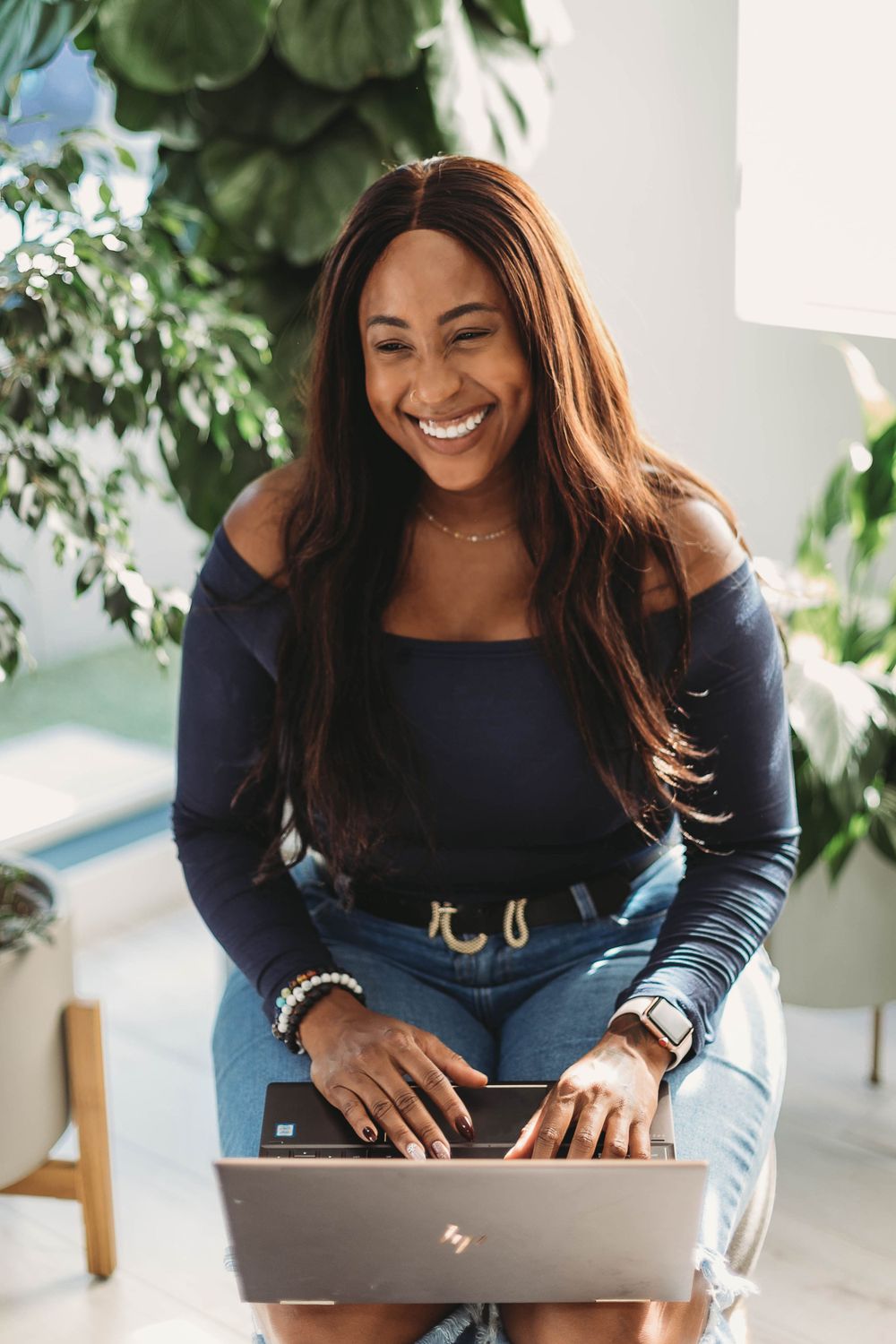 Smiling woman in navy off-shoulder top works on laptop in bright indoor setting with greenery in background.