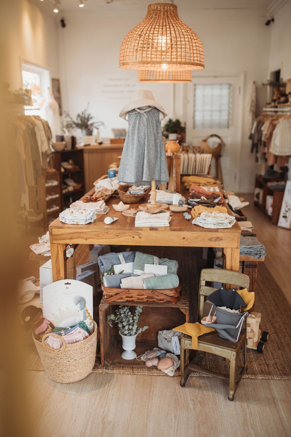 Cozy boutique interior with woven pendant lamp over wooden display table featuring baby clothes and home decor items.