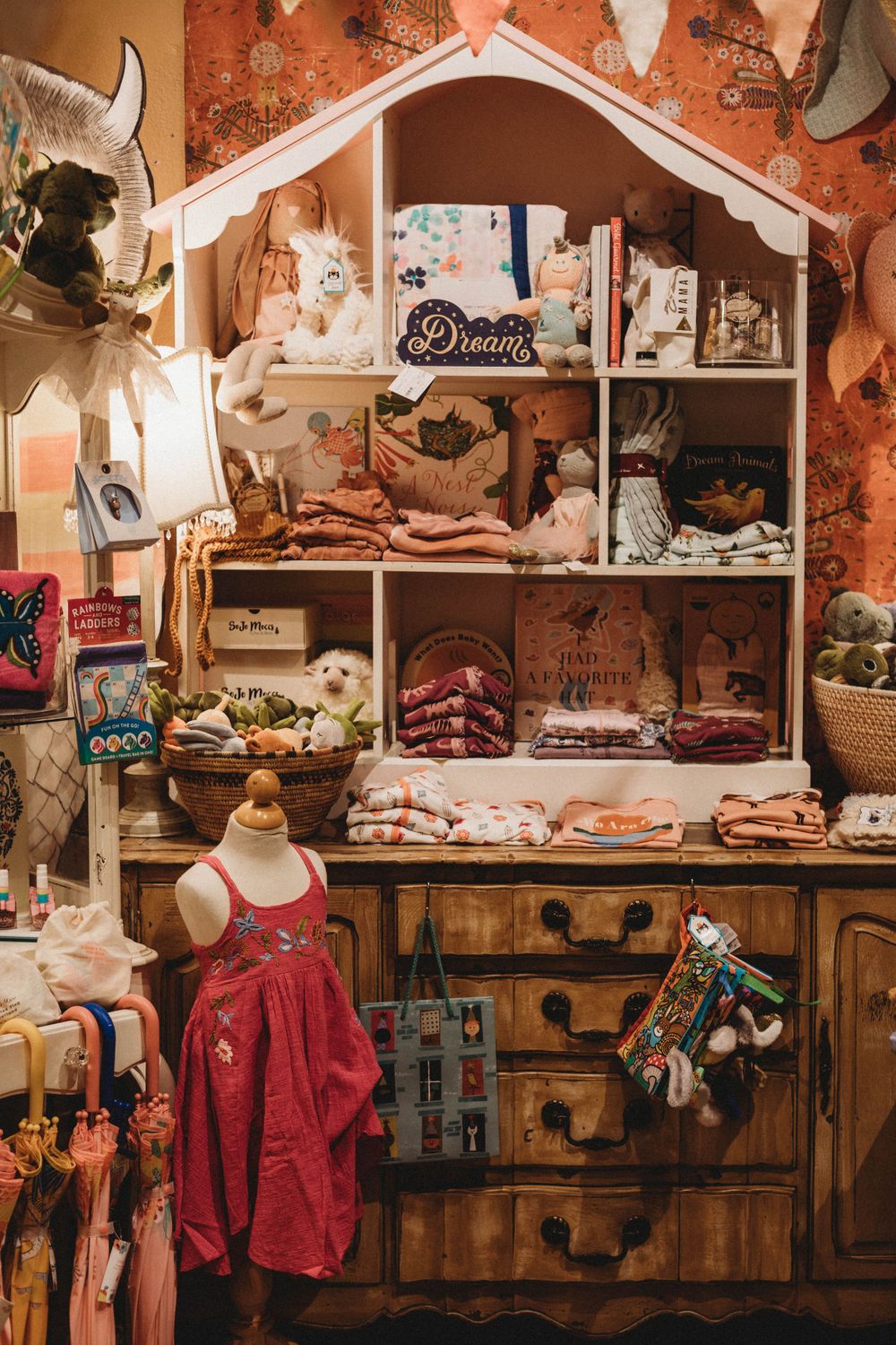 Vintage children's boutique display showing a wooden dollhouse shelf filled with toys, clothes and decorative items.