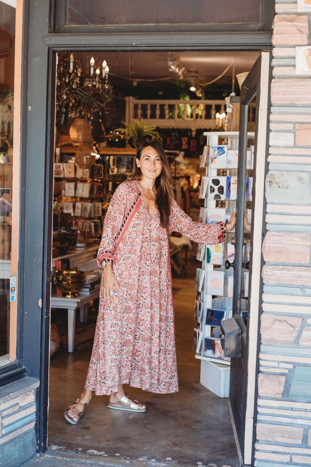A retail shop entrance displaying postcards and merchandise with a floral maxi dress in view.