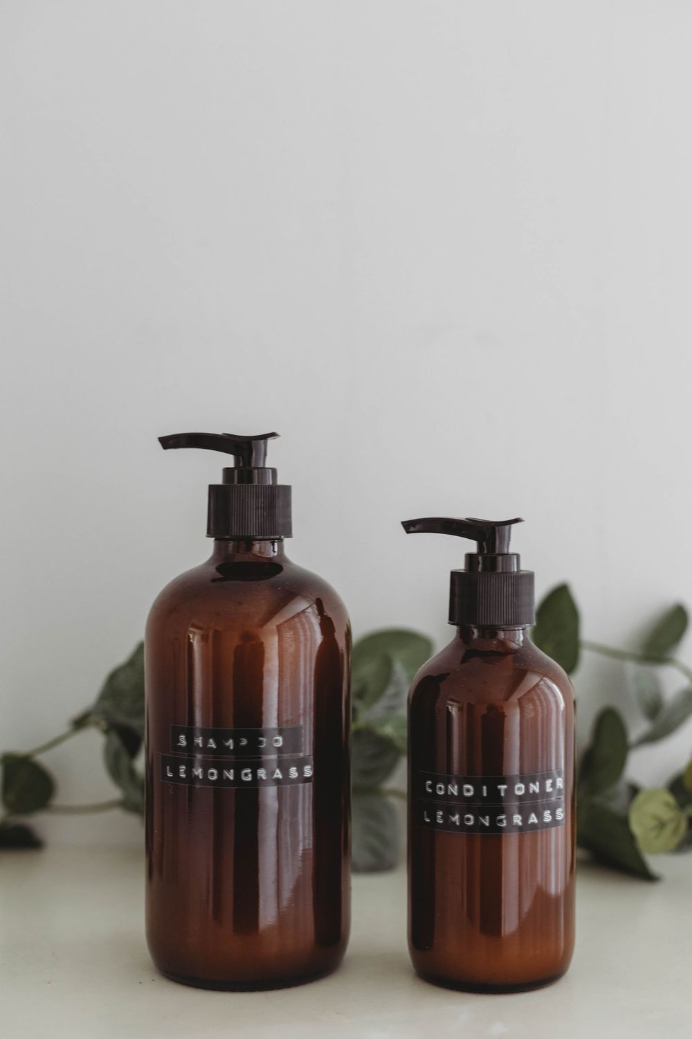 Two amber glass soap dispensers with black pumps and minimal labels sit against a white backdrop with green foliage.