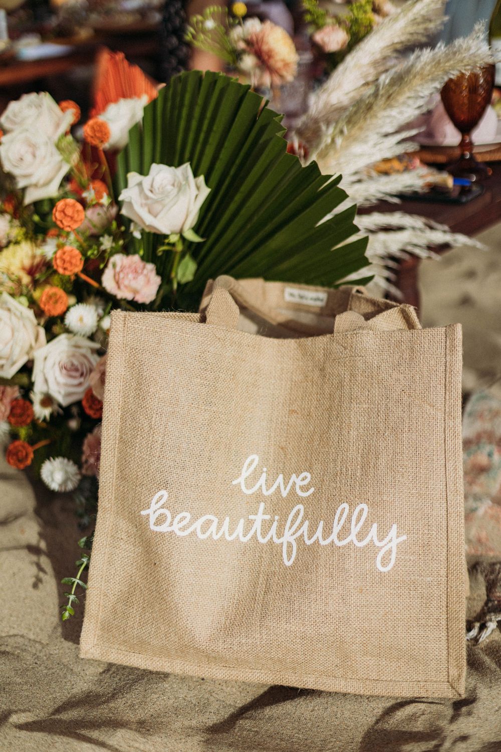 A jute tote bag with 'live beautifully' text displayed among floral arrangements with roses and palm leaves.