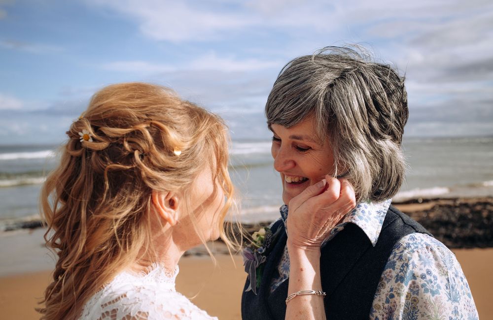 Two women share a joyful moment on a beach, one in a wedding dress, smiling at each other on a cloudy day.