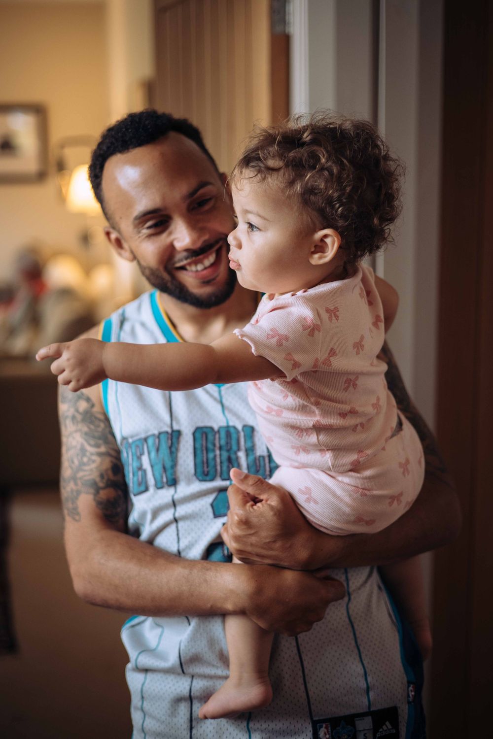 A parent in a blue jersey holds and plays with a baby wearing a light pink outfit in a warm indoor setting.