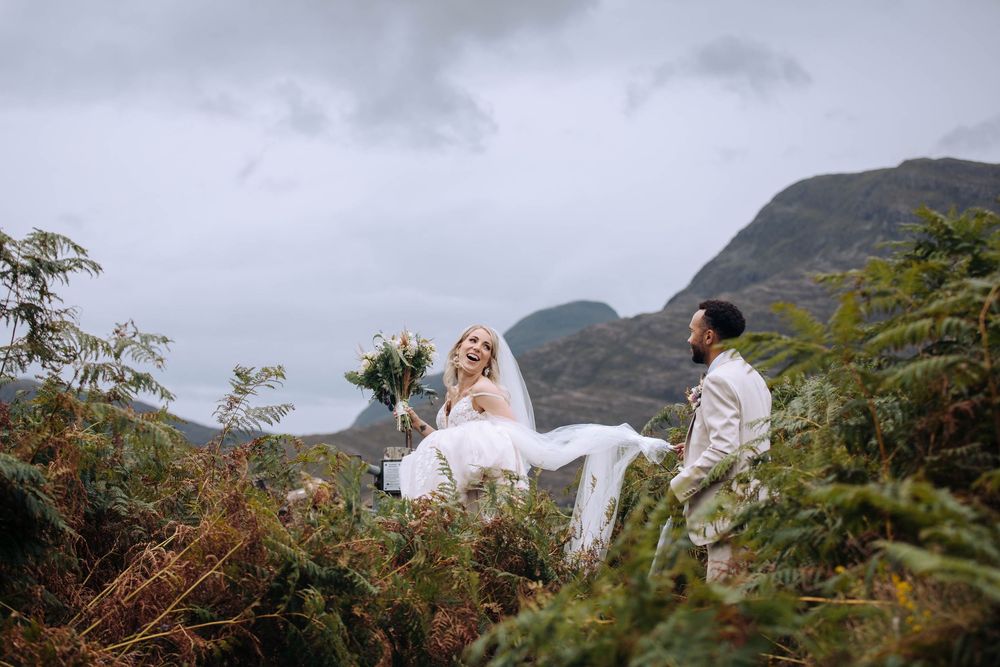 A couple in white attire sits together on a hillside surrounded by lush greenery and mountains in the background.