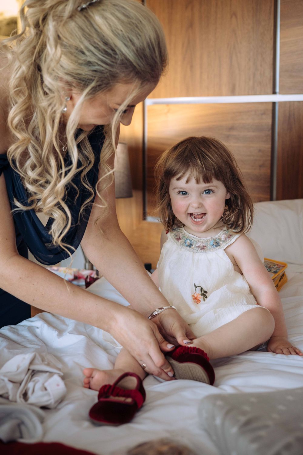 A tender moment captured as a woman helps a smiling toddler put on red shoes while sitting on a bed.