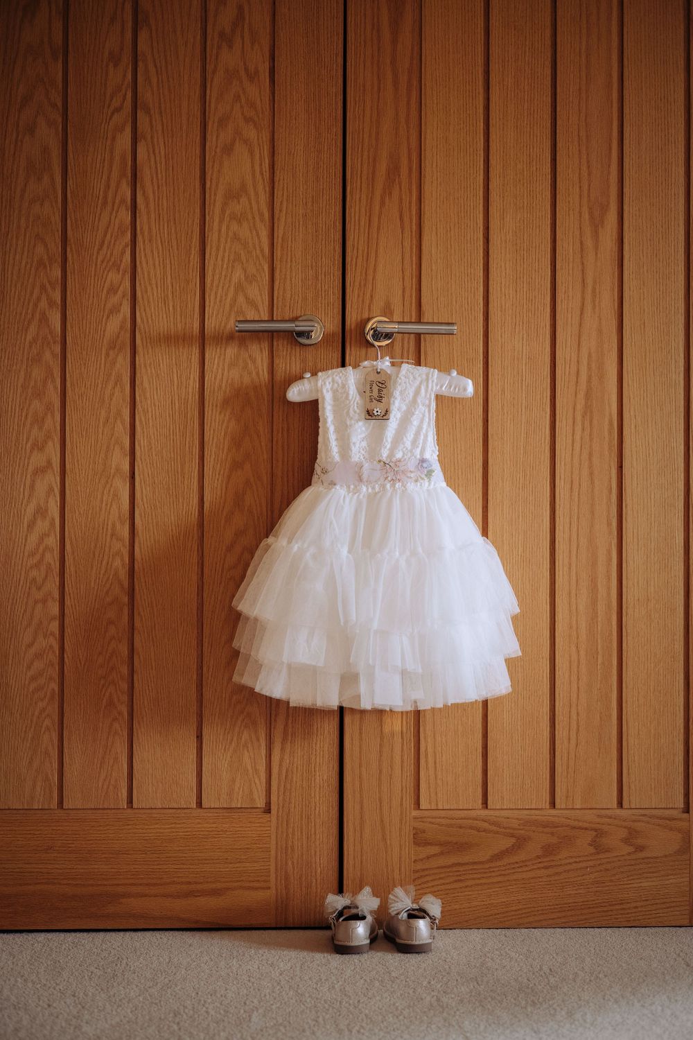 White tiered flower girl dress on hanger displayed against wooden wardrobe doors with matching shoes below.