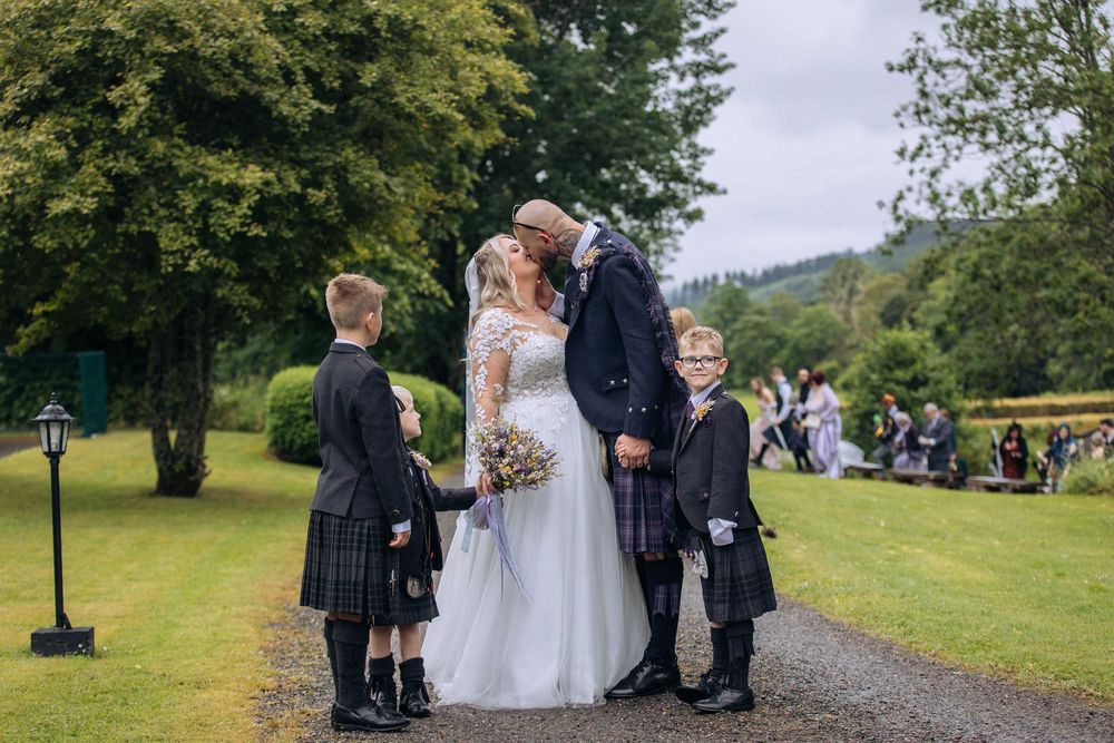 Bride and groom kissing at outdoor wedding ceremony with two boys in kilts standing beside them in a Scottish garden.