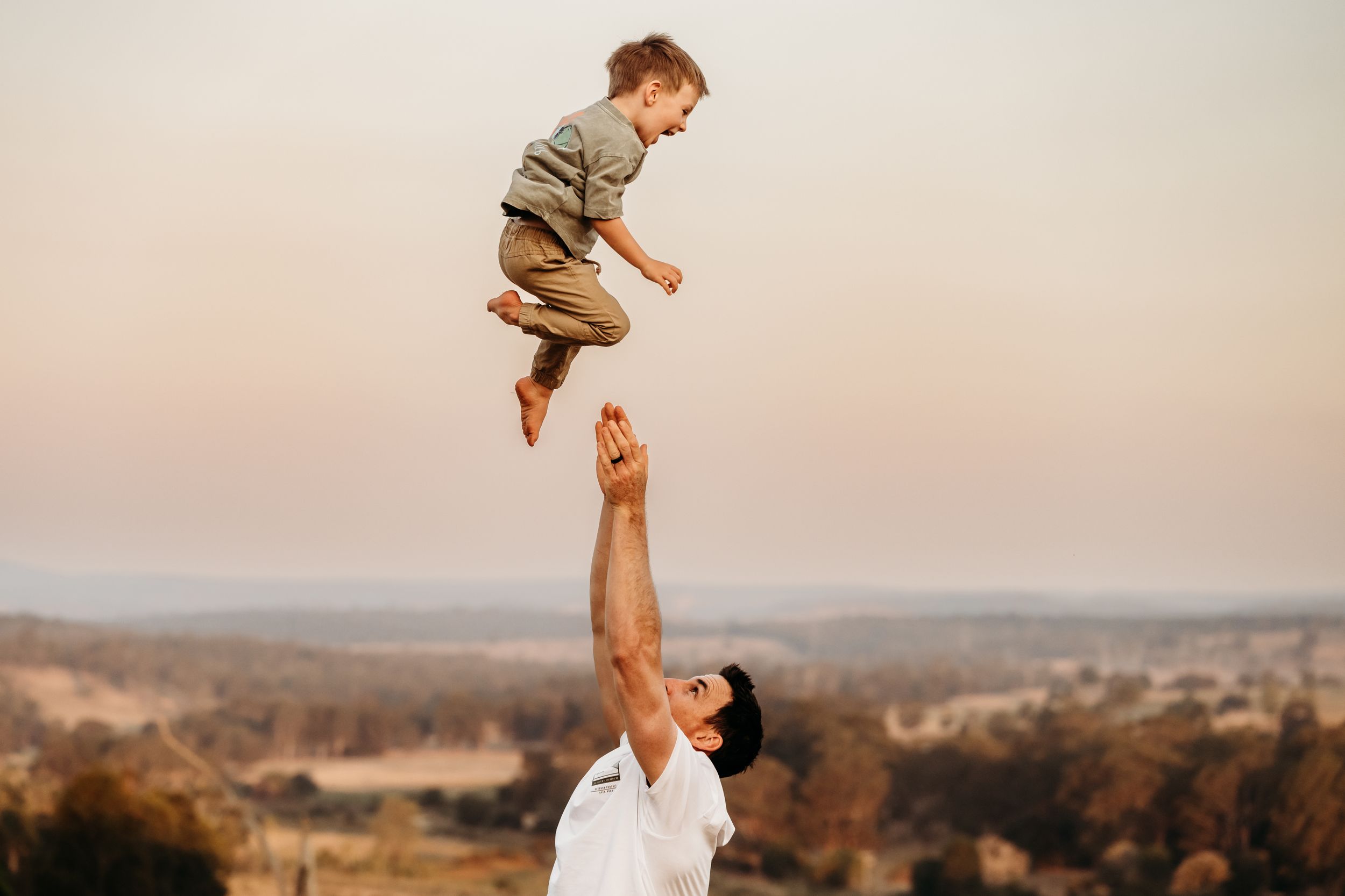 A parent playfully lifts a baby into the air against a scenic mountain landscape during golden hour.
