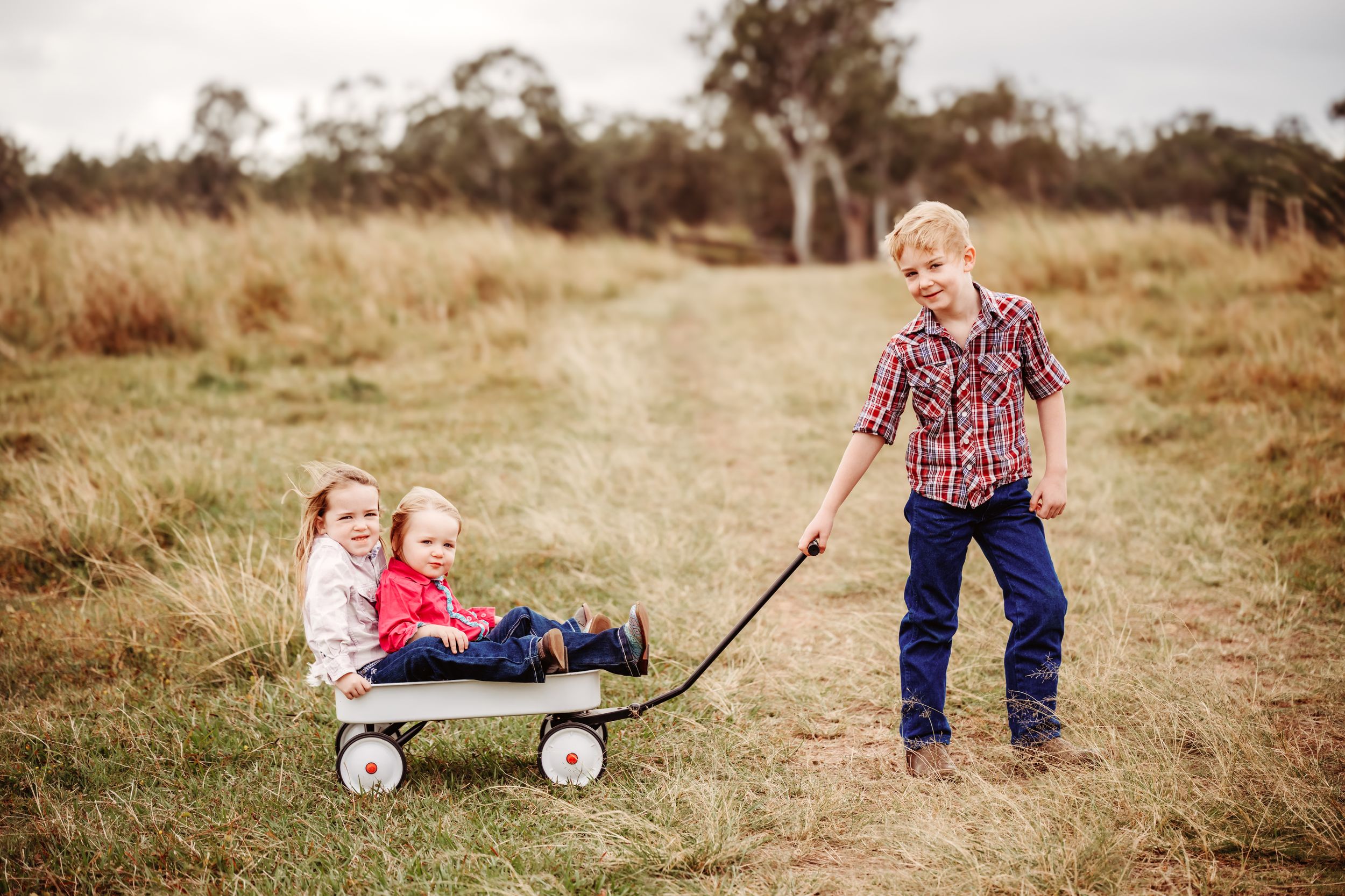 Boy pulling two young girls in a white wagon along a grassy field path outdoors.