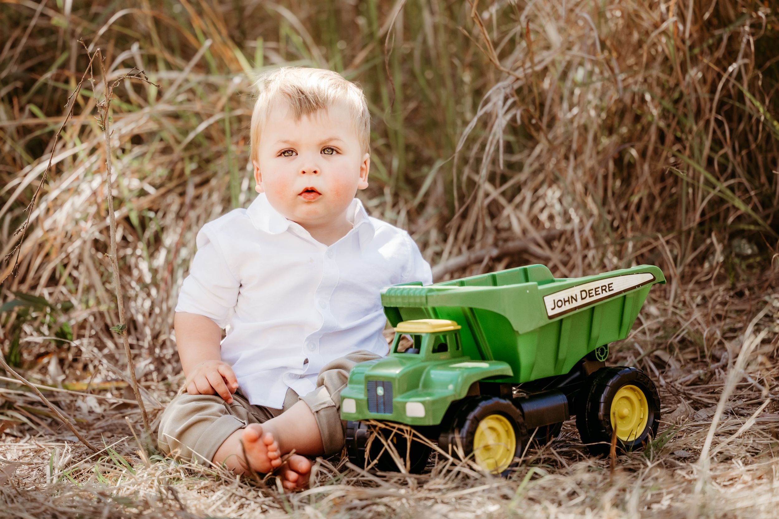 Toddler boy in white shirt sitting outdoors in tall grass playing with a green John Deere toy dump truck.