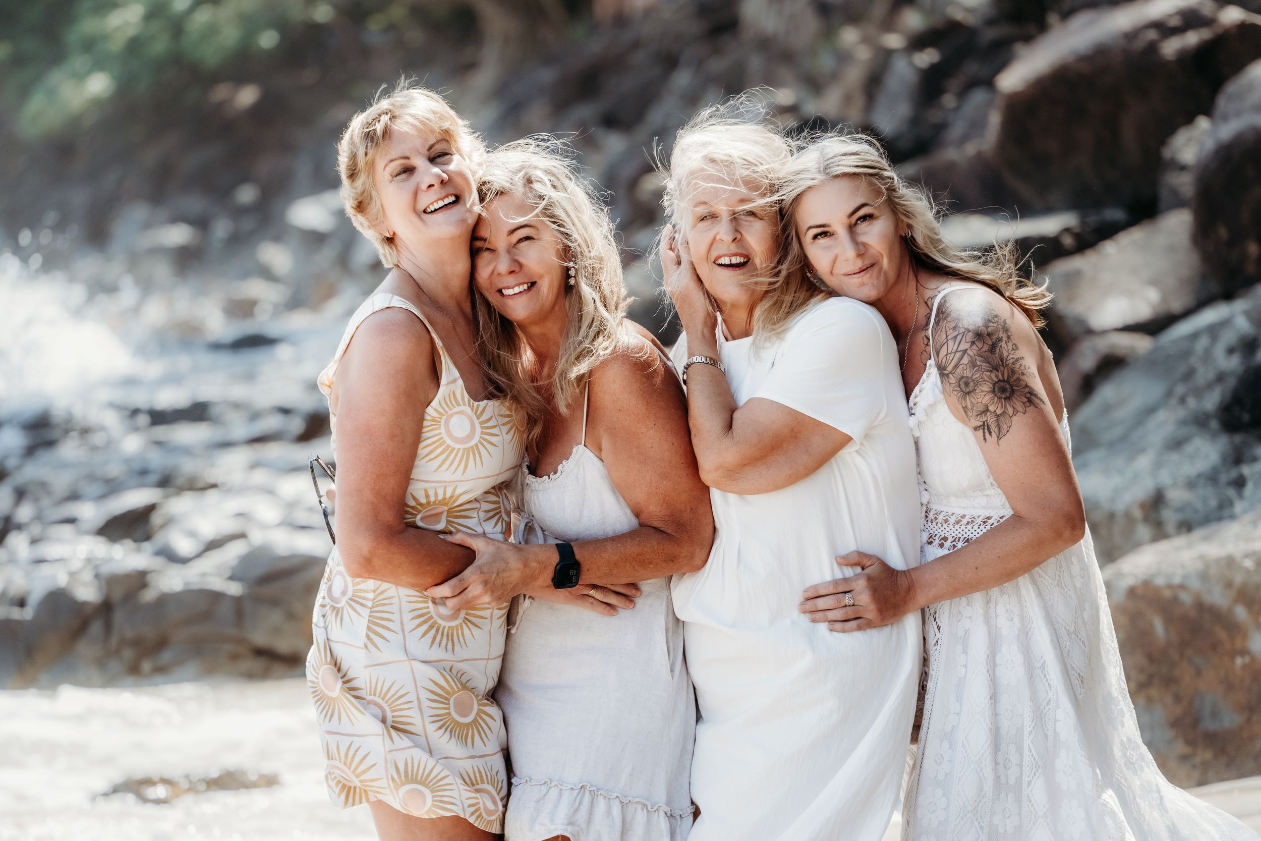 Four people in white dresses share a joyful moment together on a rocky beach with ocean spray in the background.