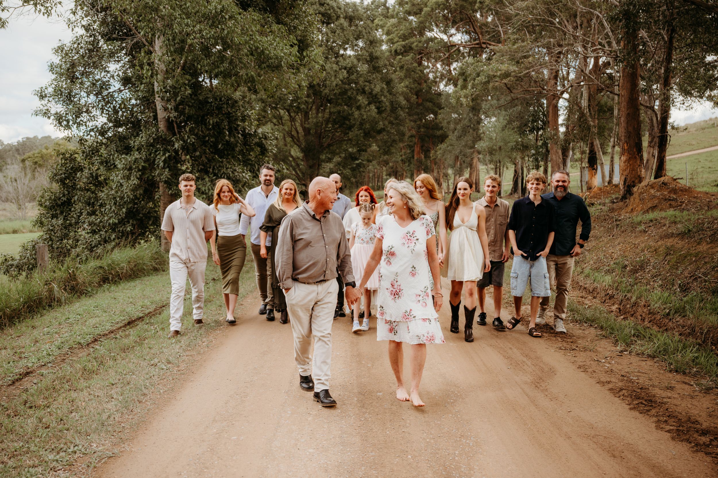 Large family group walking together on a rural dirt road surrounded by lush green trees and rolling hills.