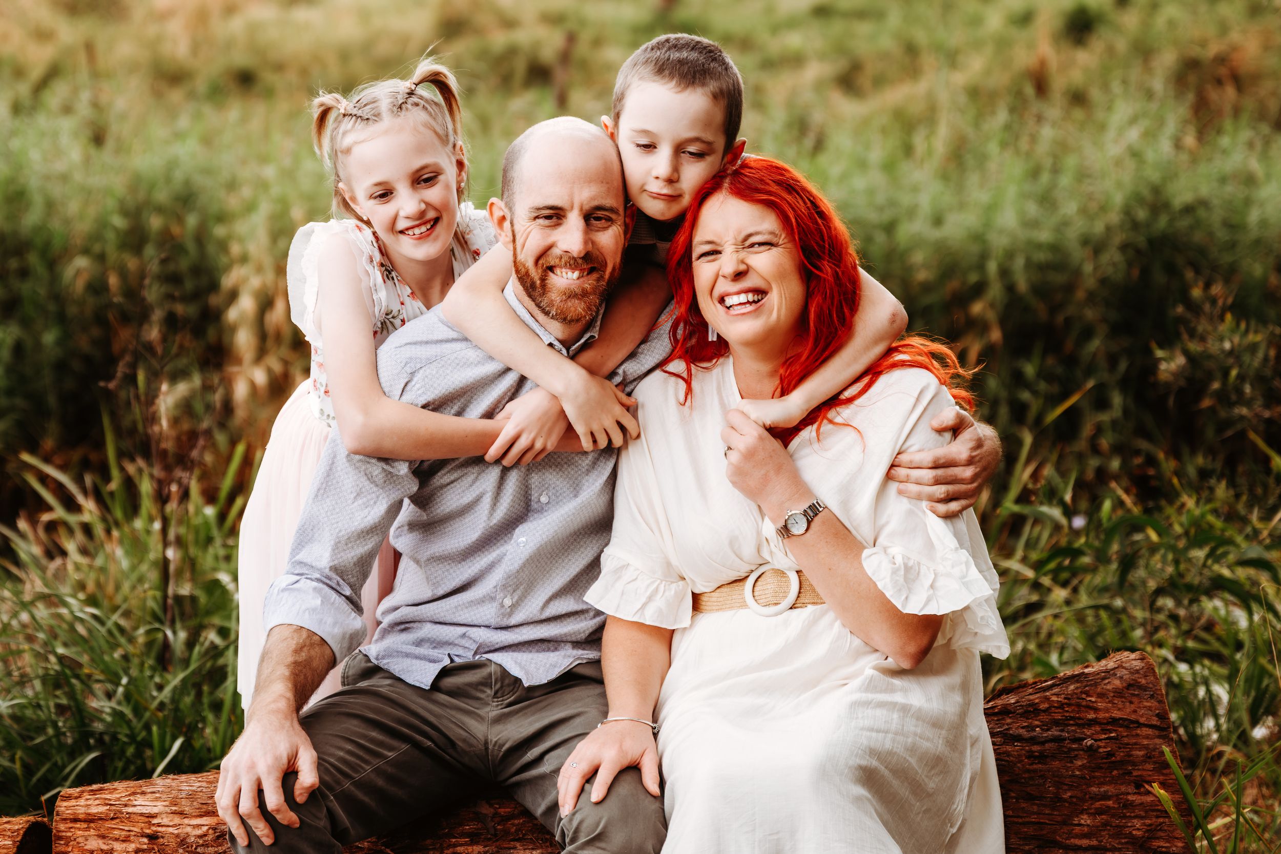 Happy family of four sitting outdoors on a log, parents and two young girls smiling in a green meadow.