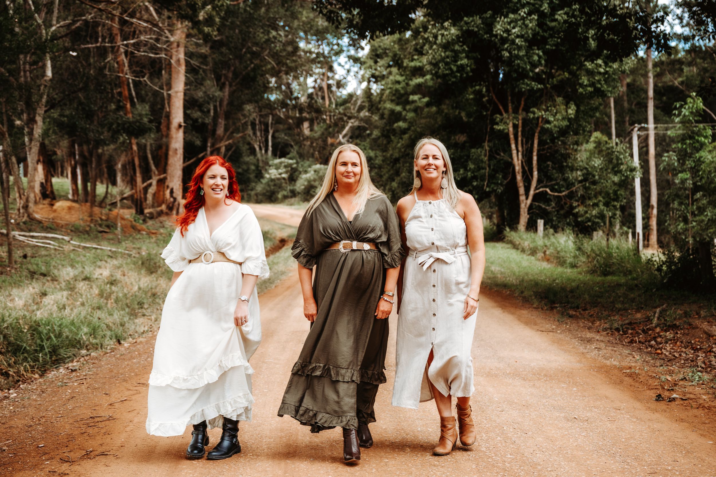 Three women in boho dresses walk together on a dirt path surrounded by lush green trees.