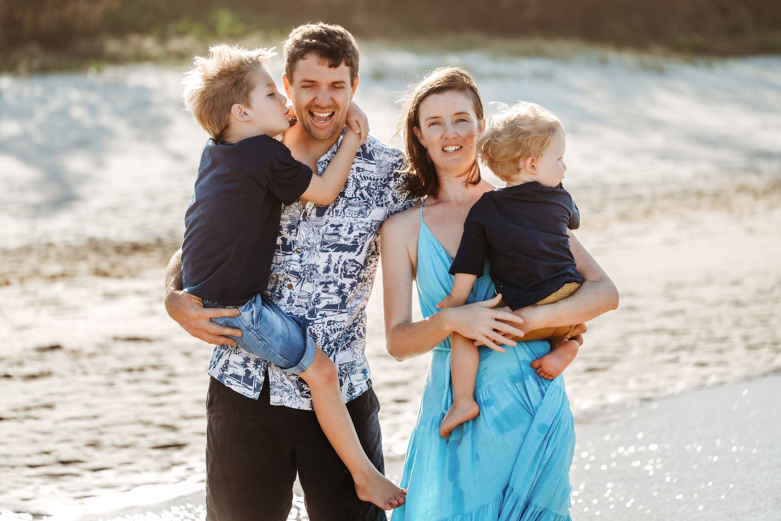 Couple in blue dress and navy shirt laughing and embracing on a sunny beach during a photo session.