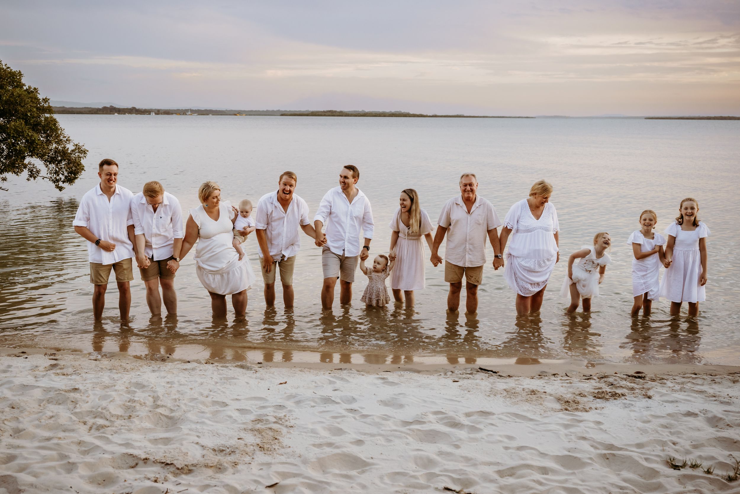 Large family group wearing white stands together in shallow ocean water at sunset.