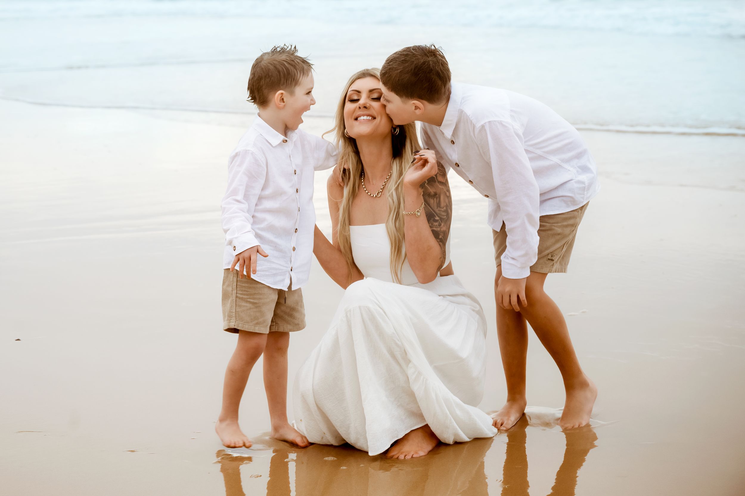 A sweet family moment captured on a white sandy beach with gentle waves in the background.