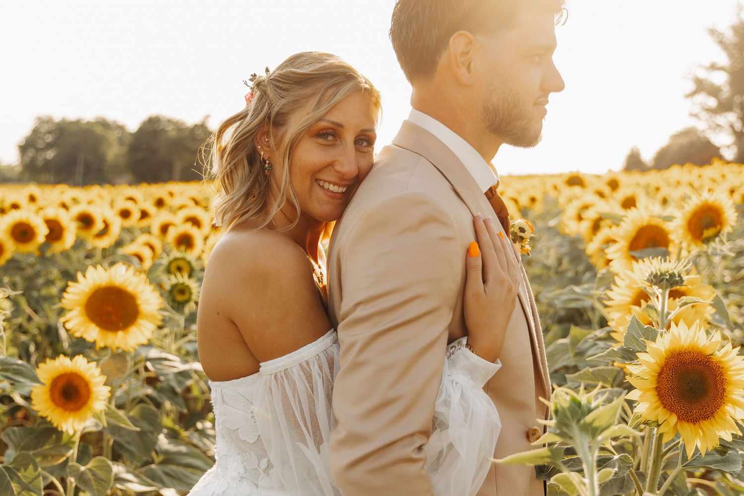 Bride smiling in sunflower field embracing groom in tan suit at golden hour.