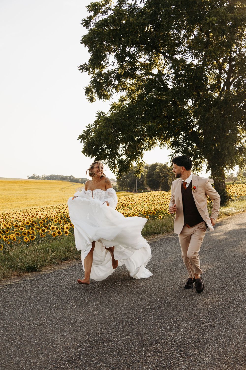 Joyful bride lifting white dress running with groom past sunflower field on country road.