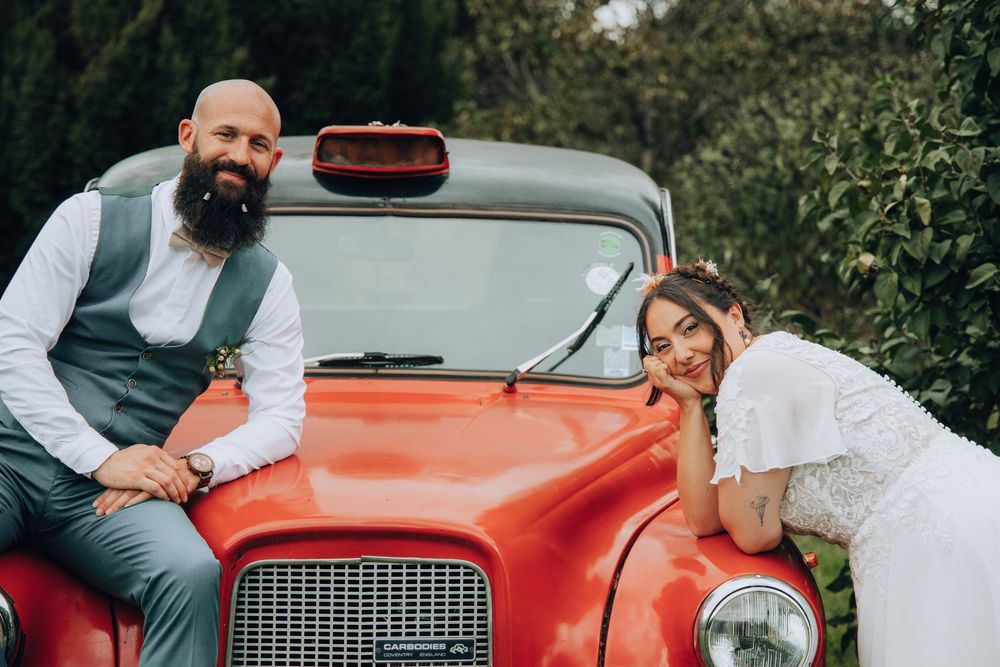 Bride and groom posing together leaning on a vintage red car on their wedding day.