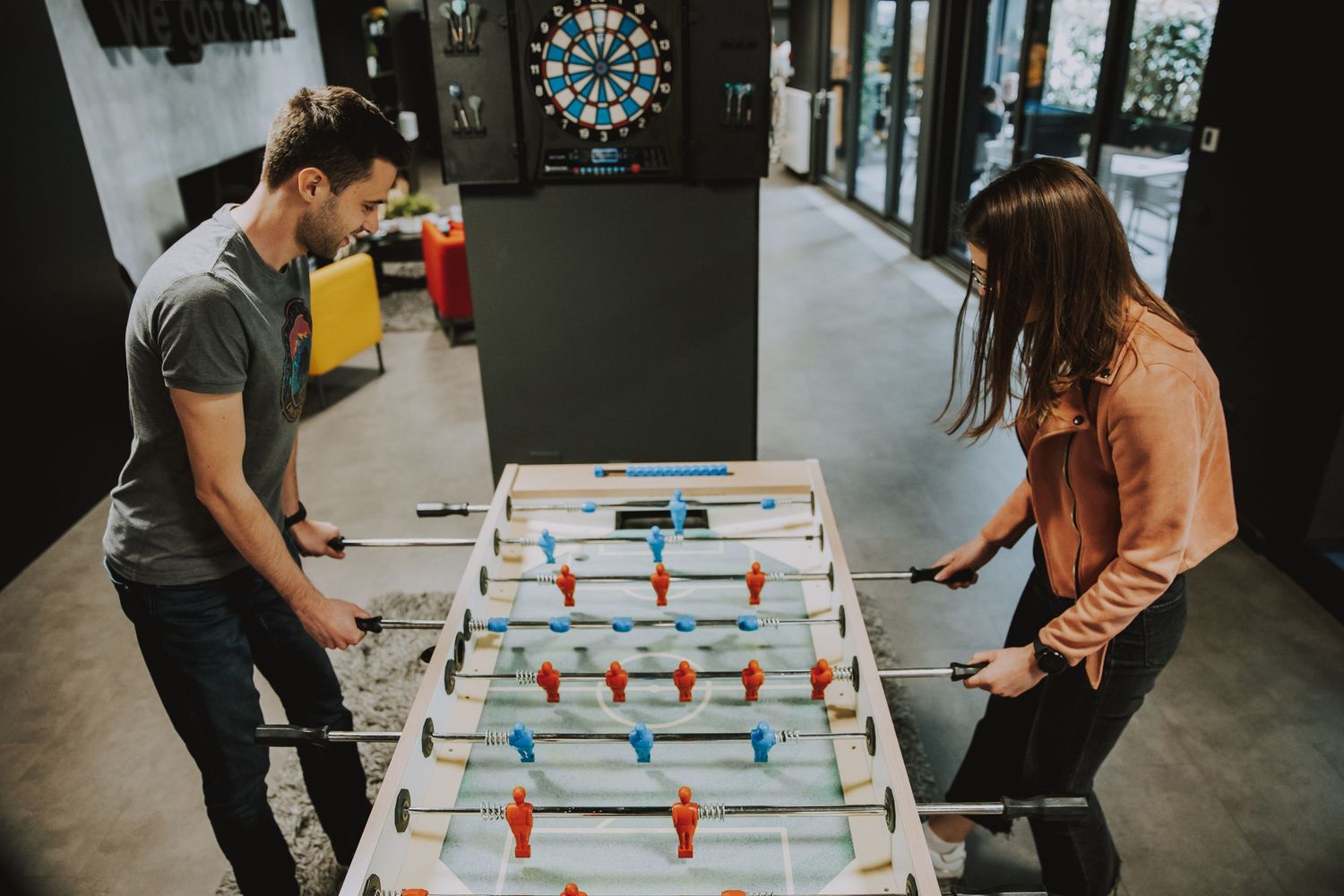 Two colleagues playing foosball table in modern office break room.