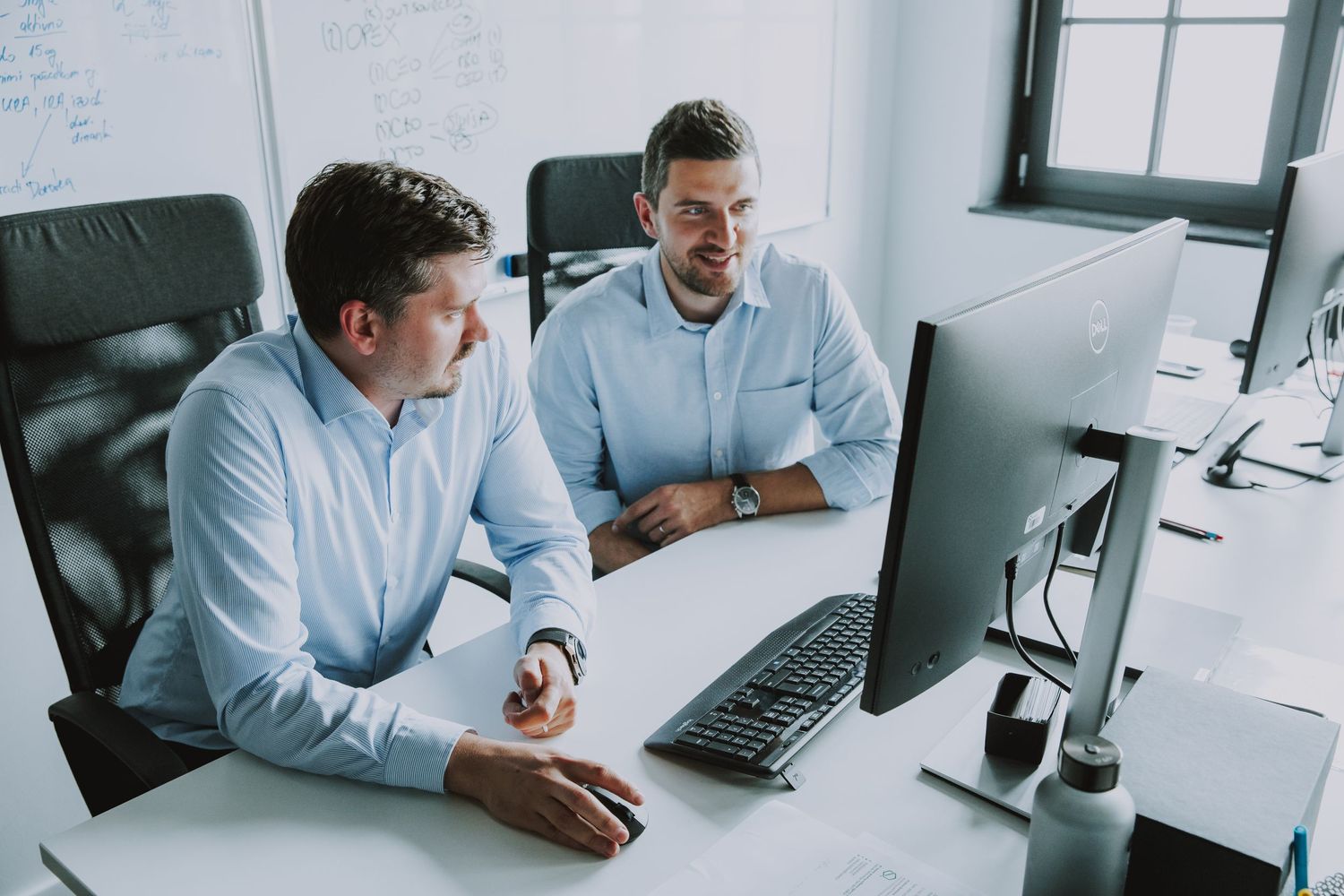 Two colleagues in light blue shirts working together at computer desk.