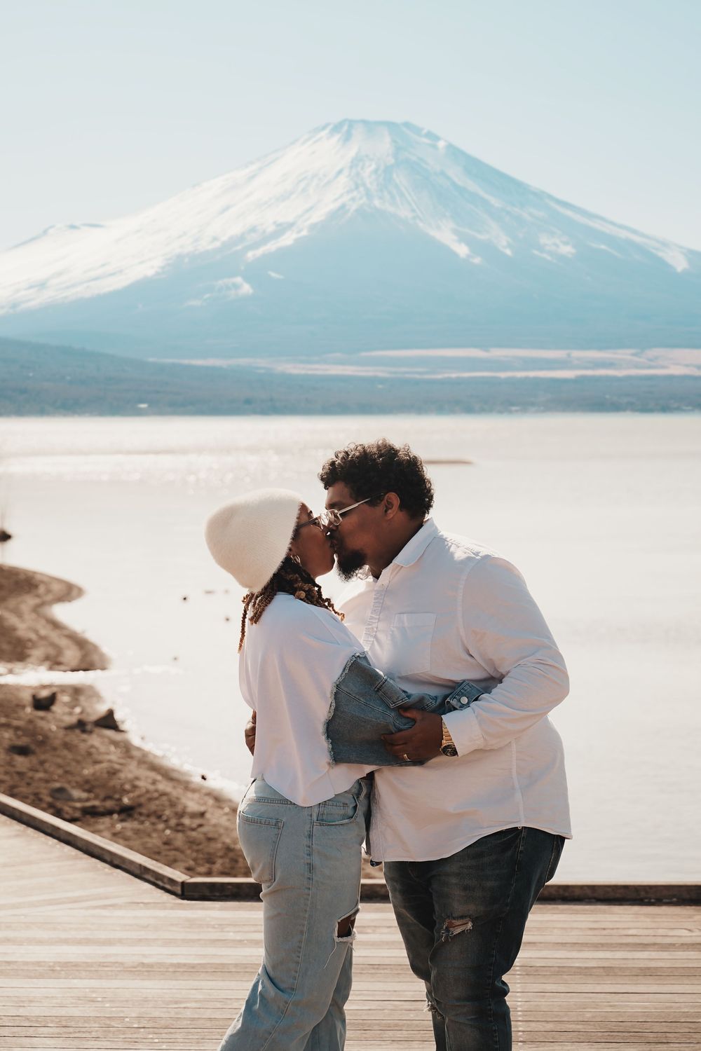 Couple embraces sweetly on a wooden boardwalk with snow-capped mountains visible in the background along a serene shoreline.