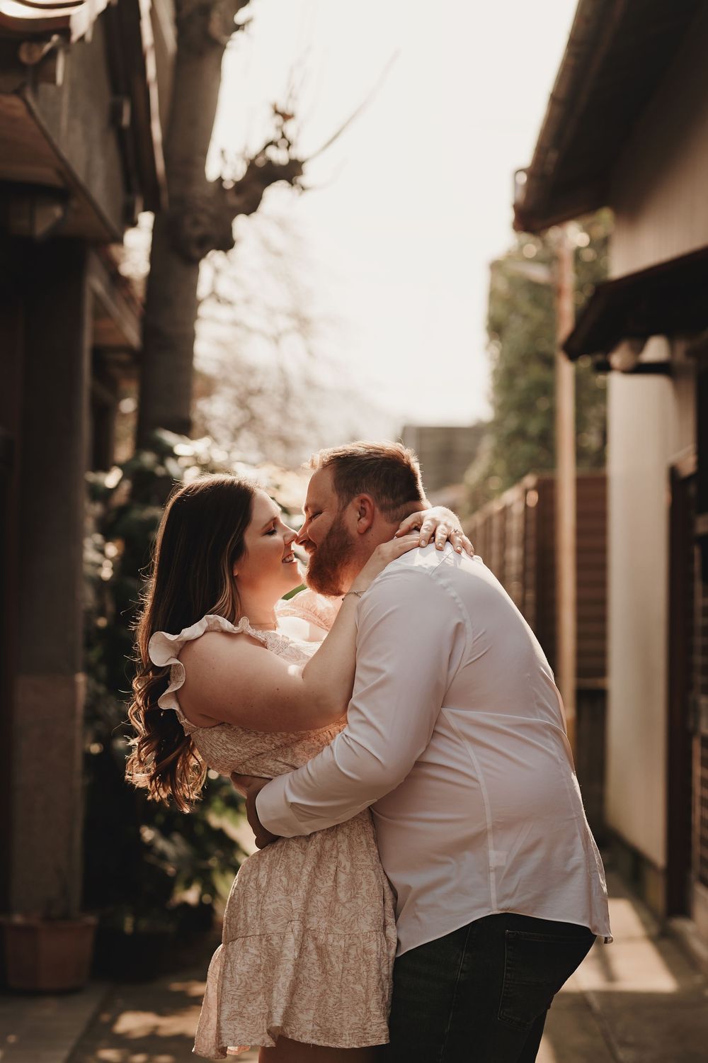 A couple in formal attire share an intimate embrace under architectural columns at sunset.
