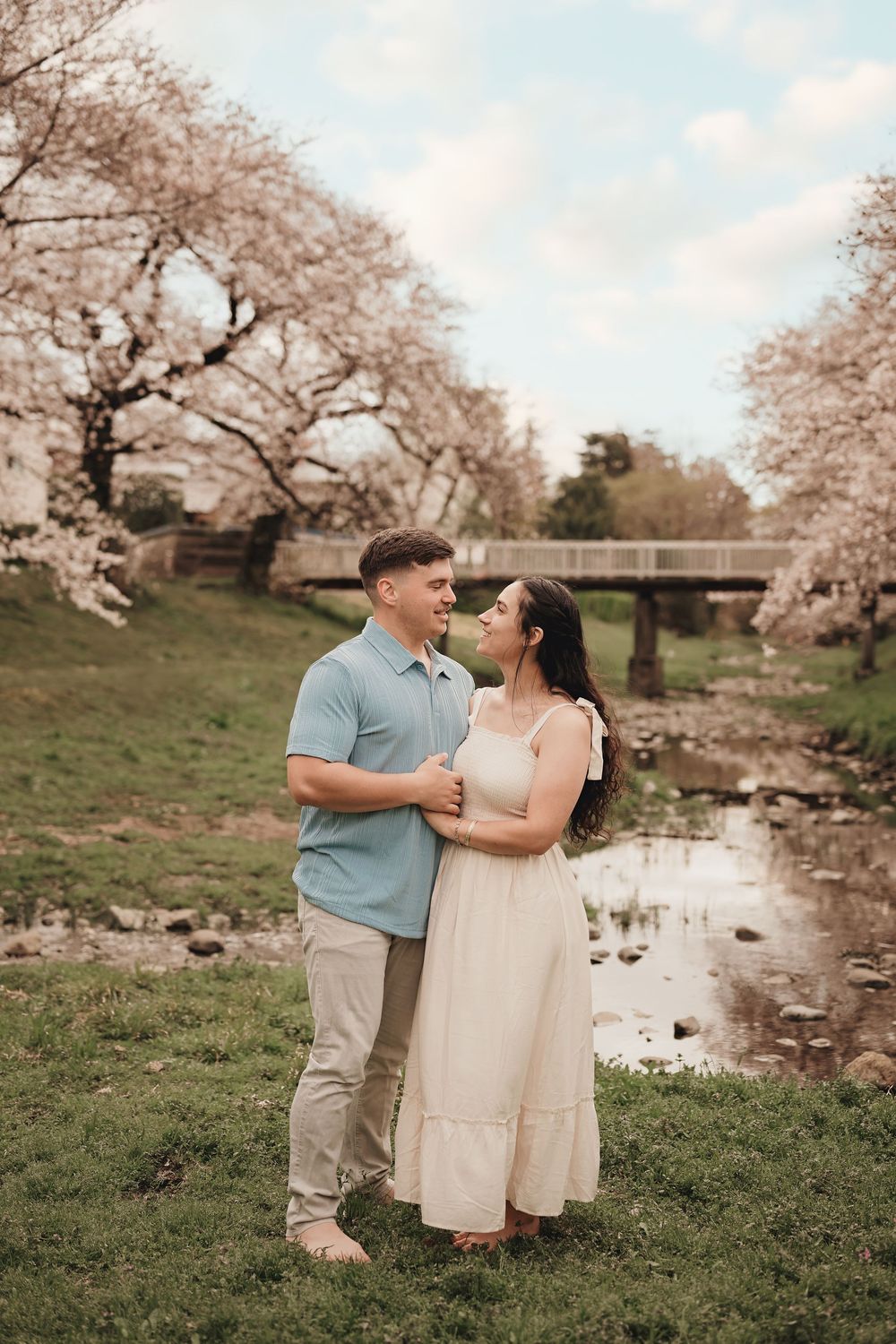 A couple embraces by a stream under blooming cherry blossom trees in a romantic springtime setting.