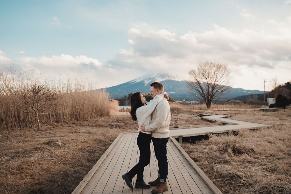 Two people embrace while standing on a wooden boardwalk with mountains and dry grass in the background at sunset.