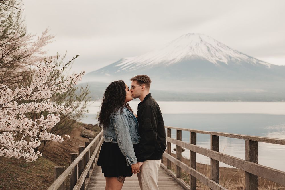 Two people share an intimate moment on a wooden walkway with snow-capped mountains in the background.